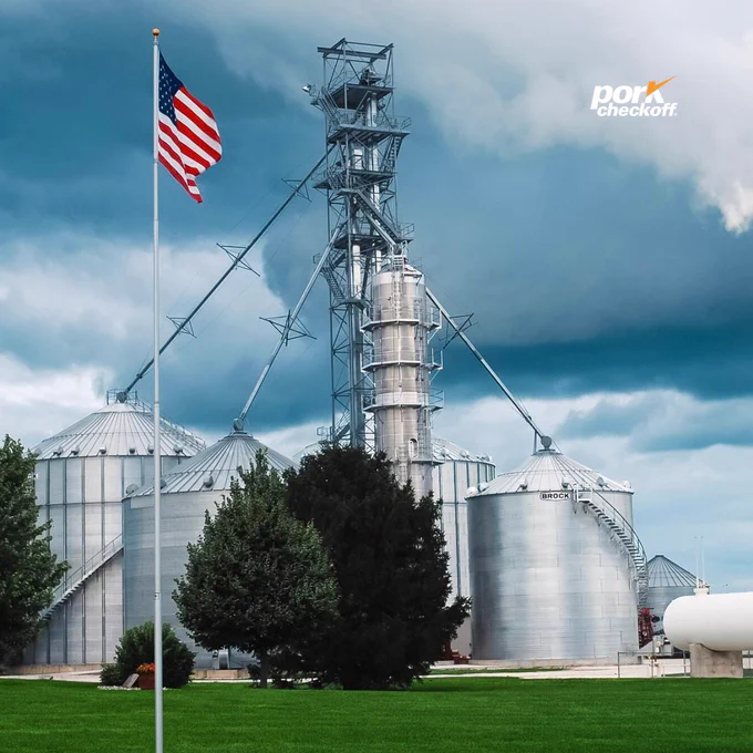 Industrial scene featuring metal grain silos and a tall structure beneath a cloudy sky. An American flag is prominently displayed on a flagpole in the foreground, set against a well-manicured lawn. The Pork Checkoff logo is visible in the top right corner.