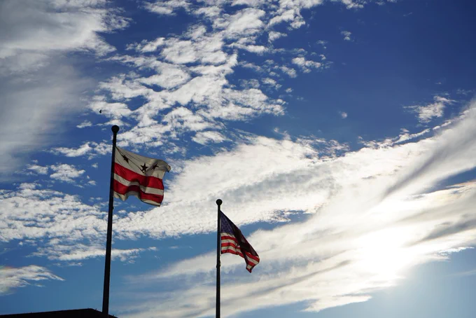 Sun is shining on the DC flag and the American flag both waving against the blue sky.
