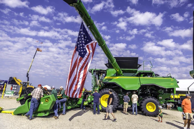 An American flag hangs from a piece of farming equipment as event attendees walk around below.