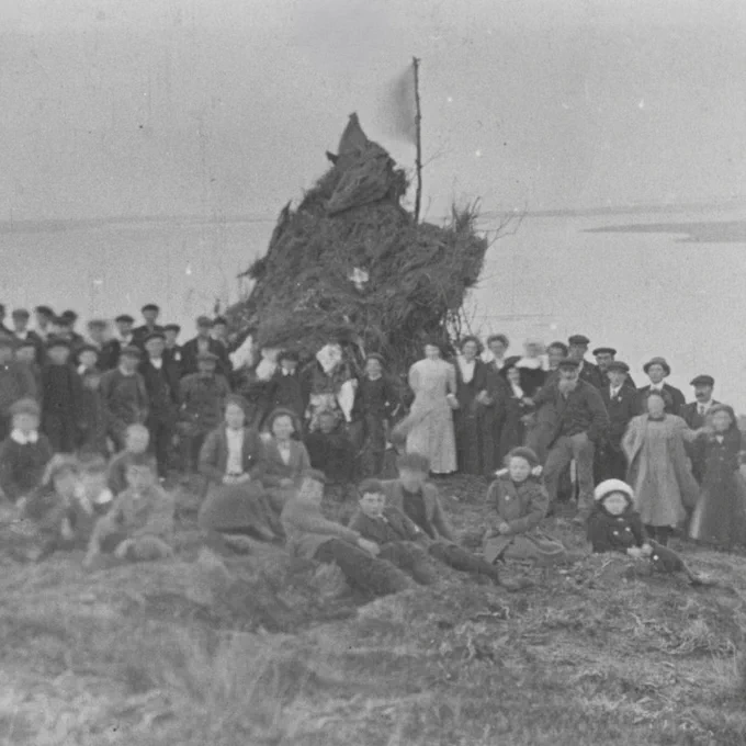 A black and white Archive photograph of a terrifying shape rising above a group of people who are looking towards the camera. It is a bonfire but looks like something more frightening.