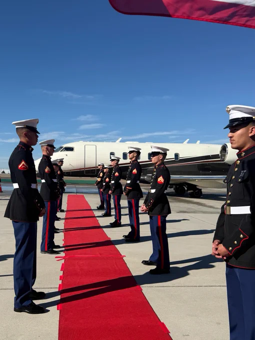Ceremonial guards in formal military attire stand in formation along a red carpet. A private jet is parked on the tarmac in the background under a clear blue sky.
