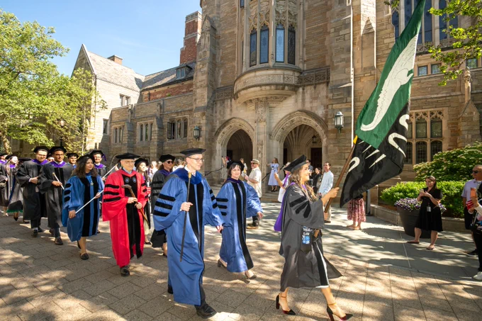 The flag bearer walks past Sterling Law Building, following by faculty and the class.
