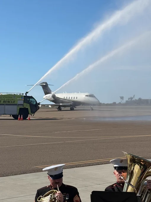 A military band is playing in the foreground as a private jet receives a water cannon salute on the tarmac. A fire rescue vehicle is visible, directing water streams over the plane. The sky is clear and blue.