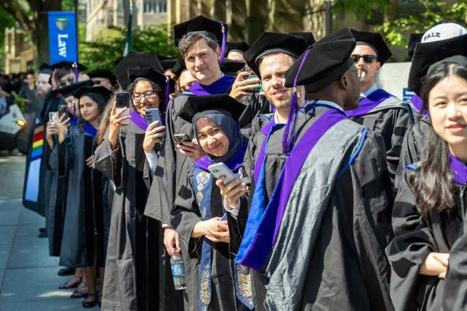A line of graduating students on campus behind a flag reading "law."