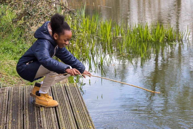 A girl stooping down over a lake with a fishing rod in her hand