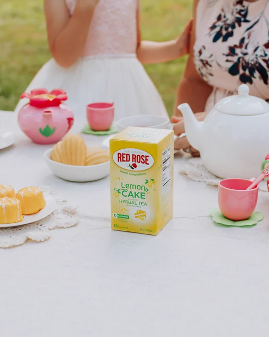 Outdoor tea party setup with a white teapot, pink floral teacups, Red Rose Lemon Cake tea, and assorted pastries on a lace tablecloth.