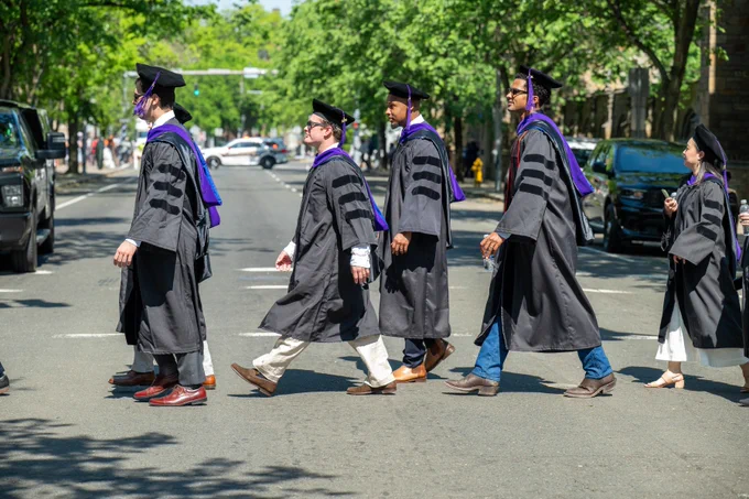 A row of graduating students in academic garb cross the street.
