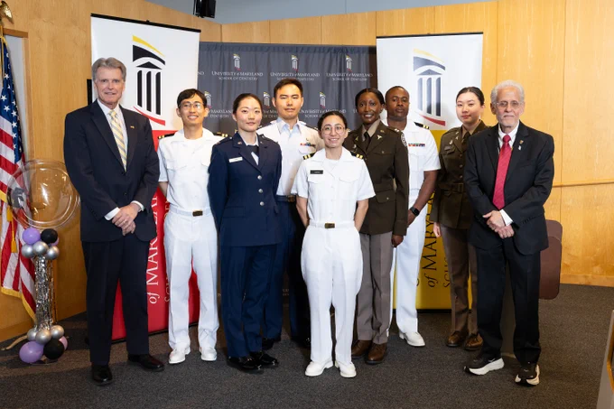 Group of military personnel and two men in suits posing together in a formal setting with banners and an American flag in the background.