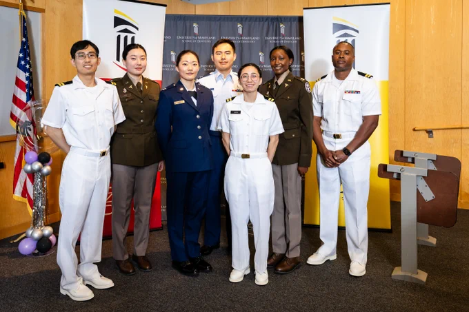 Group of six military personnel in formal uniforms posing indoors with flags and banners in the background.