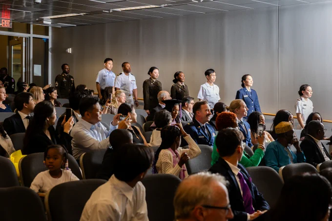 Audience seated in a conference room watching a group of uniformed individuals standing in a line near the wall.