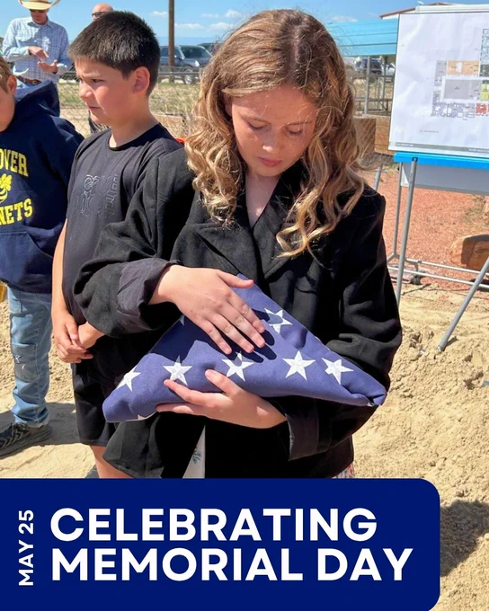 A person holding a folded American flag participates in a Memorial Day event at the Hoover Dam site. A small group is gathered, and a banner reads "Celebrating Memorial Day" with the date May 25.