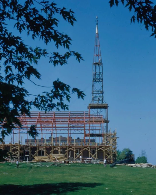 A side-view photo of the Memorial Chapel's frame under construction, with leafy branches framing the shot against a blue sky