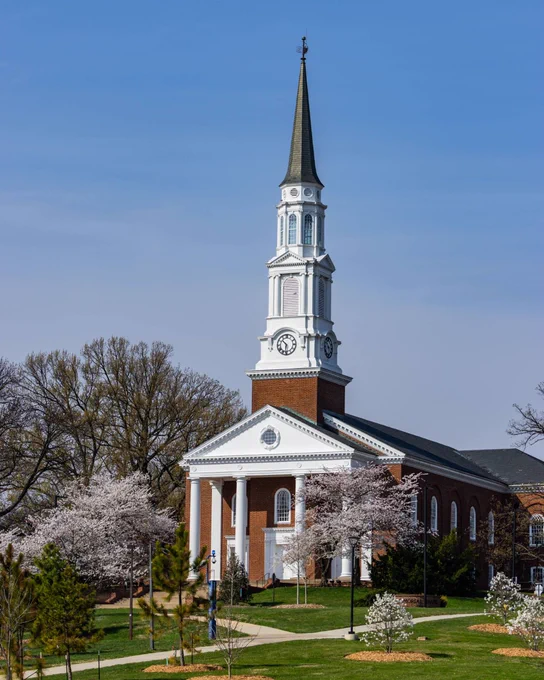 A photo of the present-day Memorial Chapel, surrounded by green and blossoming trees, underneath a pale blue sky