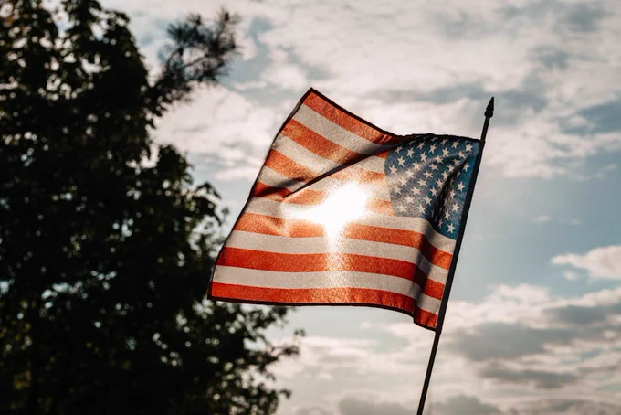 A waving American flag with sunlight streaming through against a backdrop of trees and cloudy sky.