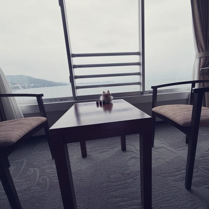 海辺のホテルの部屋の窓際の机とその奥の窓を撮った写真。窓の向こうには曇り空と海。机にはなにか人形が置いてある