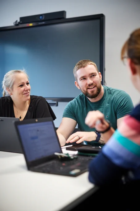 Three people are having a discussion around a table with laptops in a meeting room. A large screen is visible in the background.