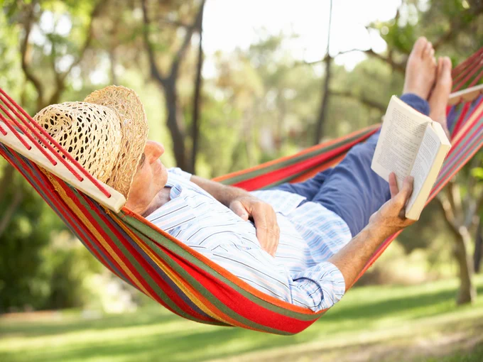 Man wearing a straw hat relaxing in a colorful hammock while reading a book in a sunny garden.