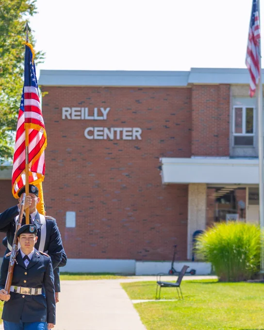 Two uniformed individuals holding an American flag in front of the Reilly Center building on a sunny day.