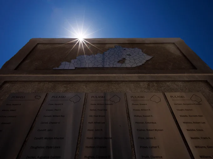A stone monument with a map of Kentucky features engraved panels listing names under headings "Powell" and "Pulaski." Sunlight shines brightly, creating a starburst effect against a clear blue sky.