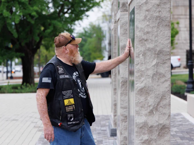 A person wearing a leather vest and cap is touching a wall with engraved text. The vest displays several patches, including one that references "Chapter 5" and displays an emblem resembling the American flag. Trees and a sidewalk are visible in the background.