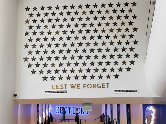 A wall displays numerous black stars above the phrase "Lest We Forget." Below, a sign reads "Kentucky." There are people in the hallway underneath the display.