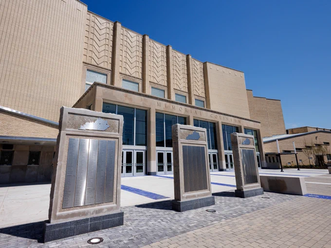 Front view of Memorial Coliseum, featuring large entrance with multiple glass doors. Three stone monuments with engraved plaques stand on a paved walkway in the foreground. The building's architecture is modern with geometric patterns. Sky is clear and blue.