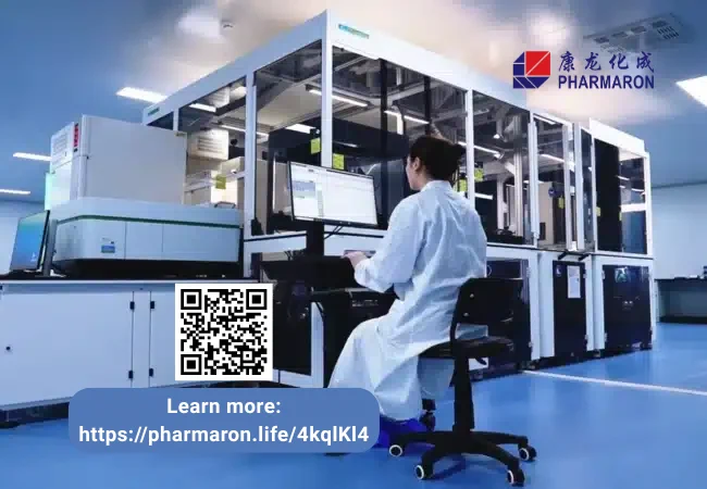 Scientist in lab coat monitors high-throughput screening system from a computer station in a clean, enclosed automation suite.