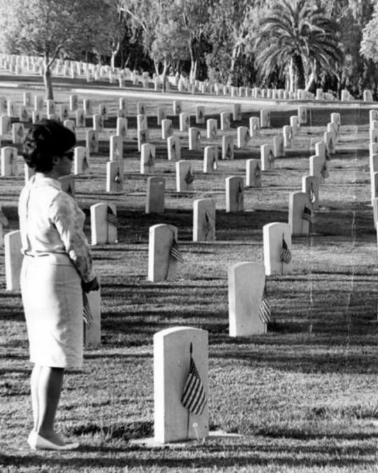 Black-and-white photo of a woman standing among rows of military headstones in a cemetery, many marked with small American flags.