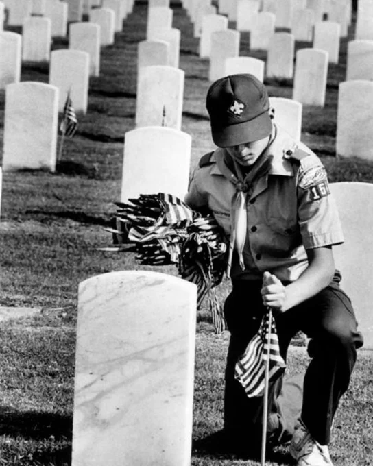 Black-and-white photo of a scout kneeling beside a military grave marker while placing small American flags in a cemetery.