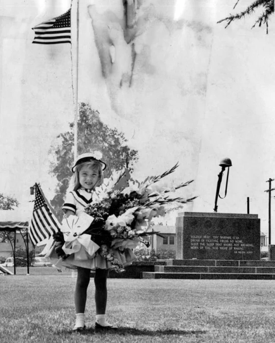 Black-and-white image of a young girl holding flowers and a small American flag near a war memorial monument.