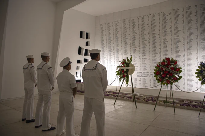 Four sailors in white uniforms stand solemnly before a large memorial wall inscribed with numerous names and adorned with floral wreaths.