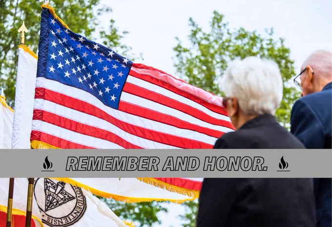 An American flag waves in the foreground, with two individuals observing. The words "Remember and Honor" are displayed on a banner across the image. Trees are visible in the background.