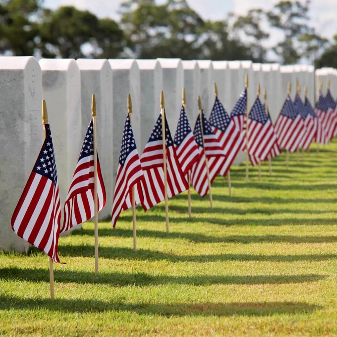 A row of white headstones in a cemetery is adorned with numerous small American flags, each placed in front of a headstone. The scene is set under a blue sky with trees in the background. 📸 ID 197590026 ©  Traci Sproule | Dreamstime