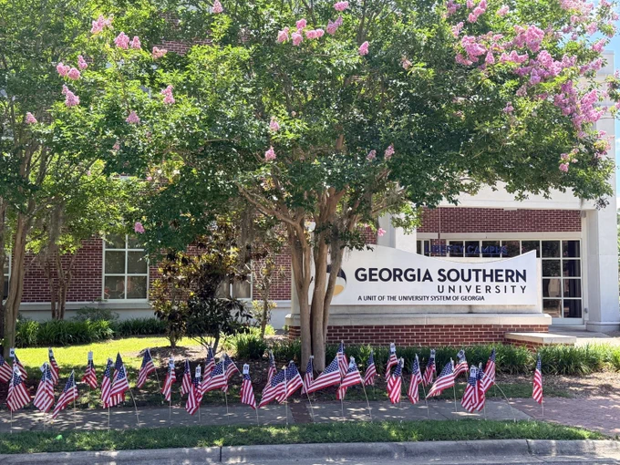 The image shows the entrance of Georgia Southern University with a sign on a brick building. The surrounding area has trees with pink flowers and numerous small American flags lined up in front of the sign.