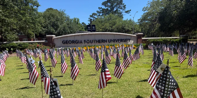 A grassy area in front of a Georgia Southern University sign is filled with numerous small American flags. Trees and a clear blue sky are in the background.