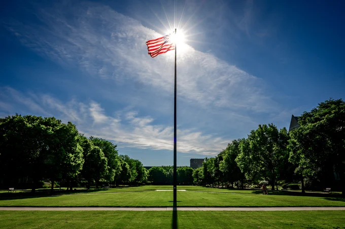A lone flagpole with an American flag stands in the center of a manicured lawn. Sunlight streams through the flag, and a row of trees lines the background under a clear blue sky.