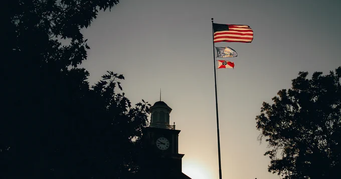 An American flag and two other flags are flying on a flagpole against a dusky sky. A large clock tower is partially visible in the background, surrounded by silhouetted trees.