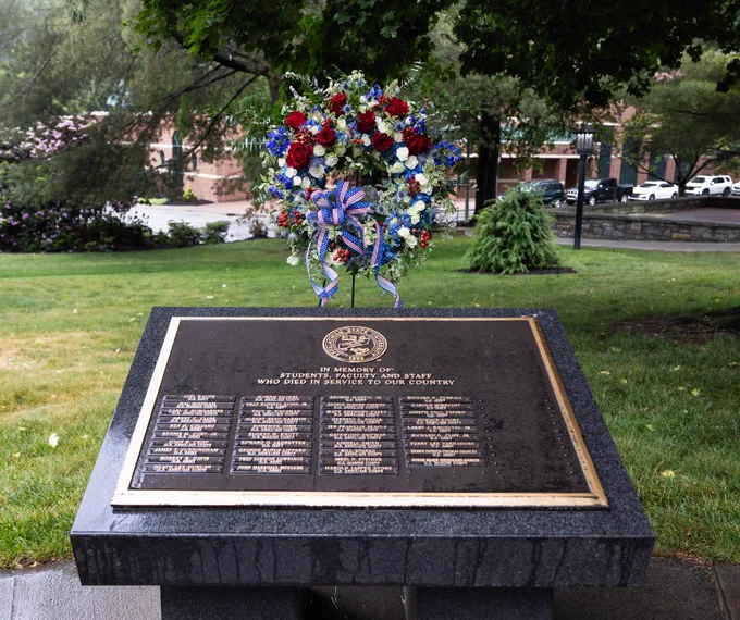 A memorial plaque set on a stone pedestal honors fallen service members. The plaque displays engraved names under an emblem. A colorful wreath with red, white, and blue flowers, along with ribbons, is mounted above the plaque. The background features a park setting with trees and buildings in an urban environment.