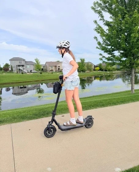 A person wearing a white helmet, white shirt, and denim shorts rides an electric scooter on a smooth path. The setting is a peaceful residential area with green grass, a tree, and a reflective pond in the background under a partly cloudy sky.