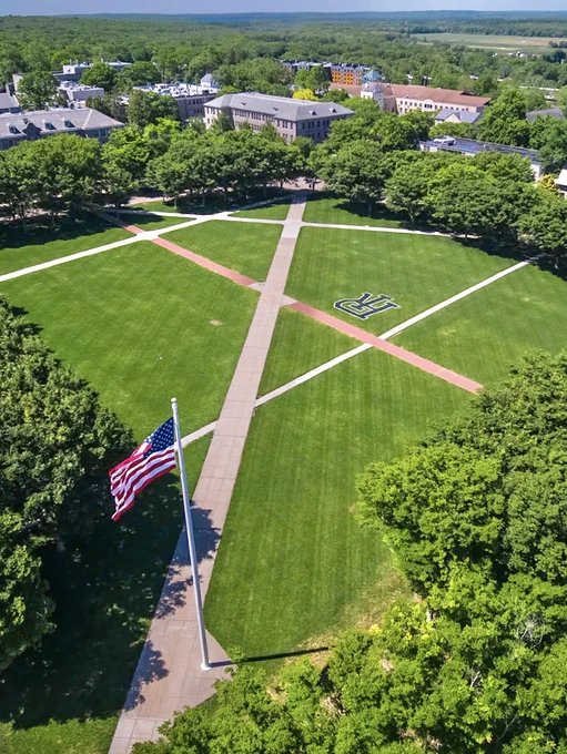 Aerial view of the URI Quad with intersecting walkways, an American flag on a pole, and a large interlocking R.I. logo on the grass surrounded by trees and campus buildings.