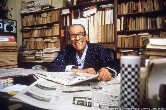 Naguib Mahfouz sitting at a desk covered in newspapers smiling. Behind him are shelves overflowing with books.