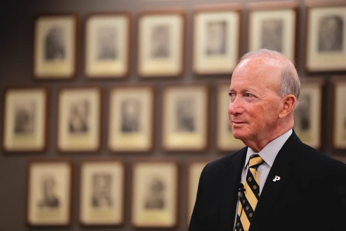 This image shows a person in a suit with a “P” lapel pin, standing in front of a wall with several framed, sepia-toned photographs.