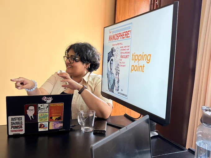 A picture of a person in front of a big monitor and a laptop. They seem to be interacting with someone in the room as their finger points towards someone. The background is a yellow wall.