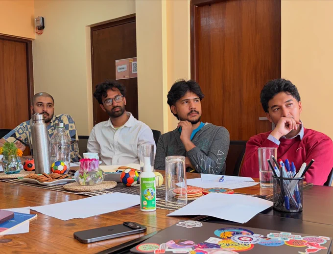 A zoomed out picture of the room where four people are sitting in the table. All of them seemed to be looking at the monitor which is in front of them. There are papers, glasses and laptops spread across the table.