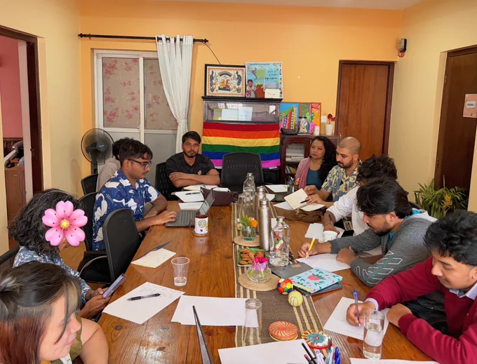 A pan view of office space of Body & Data where people are sitting across the table while they look at the screen. The backdrop is a yellow room with some curtains on the background. Some people are writing on a paper or their laptops.