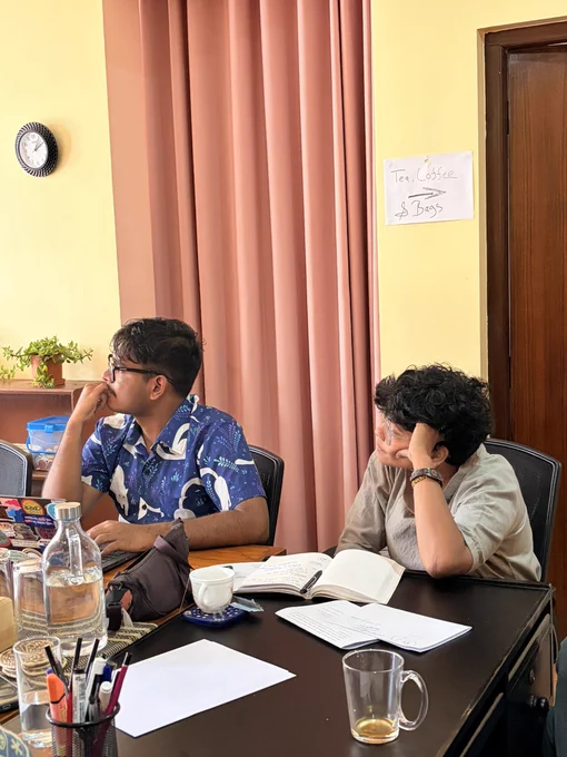 Two people rest their arms on the table and they are looking on their left on the screen. There are cups, papers, and some stationeries on the table.
