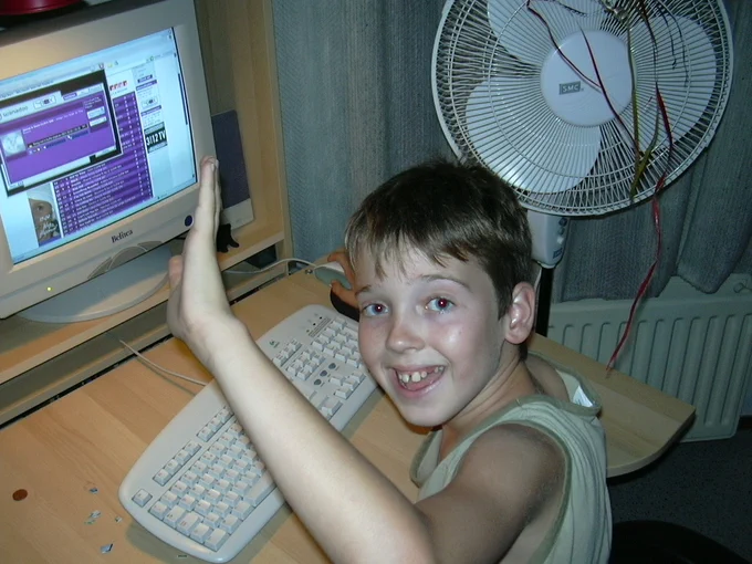 A smiling boy with short hair sits at a computer desk, waving at the camera, with a screen displaying a colorful webpage and a fan in the background.