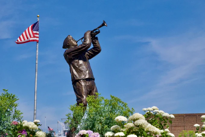 A bronze statue of a person playing a trumpet is surrounded by lush greenery and flowers. An American flag is visible in the background against a blue sky.