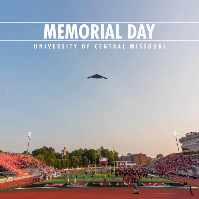 Wide view of a football stadium at the University of Central Missouri filled with people in red attire. A B-2 Spirit stealth bomber flies overhead against a clear sky. The text "Memorial Day" and "University of Central Missouri" is displayed at the top.