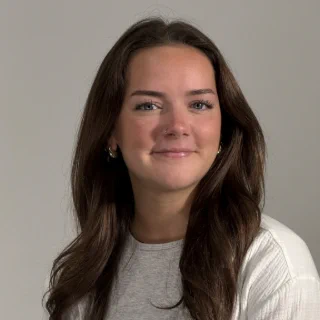 A close up headshot of Julianna, wearing a white top in front of a gray background.